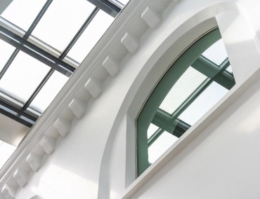 architektur_hrg Blick nach oben auf weiße Wand mit grünem Fenster und Glasdach mit Stahlträgern.