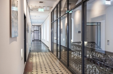 hrg_flur Langer Flur mit gemustertem Boden, weißen Wänden und großen Glasfenstern mit schwarzem Metallrahmen auf der rechten Seite.