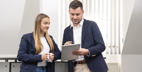 team_gespraech Zwei Personen in Businesskleidung stehen nebeneinander, eine hält eine Tasse, die andere ein Notizbuch.