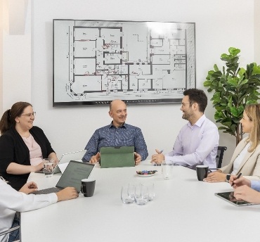team_runder_tisch Sechs Personen sitzen an einem weißen Konferenztisch, im Hintergrund ein großer Plan eines Gebäudengrundrisses an der Wand, rechts eine grüne Pflanze.