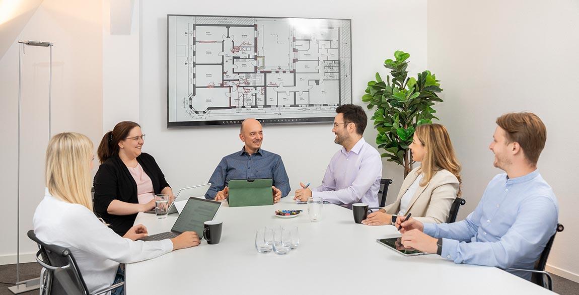Sechs Personen sitzen an einem Konferenztisch, vor einem Wandbild mit einem Gebäudegrundriss, mit Laptops und Kaffeetassen.