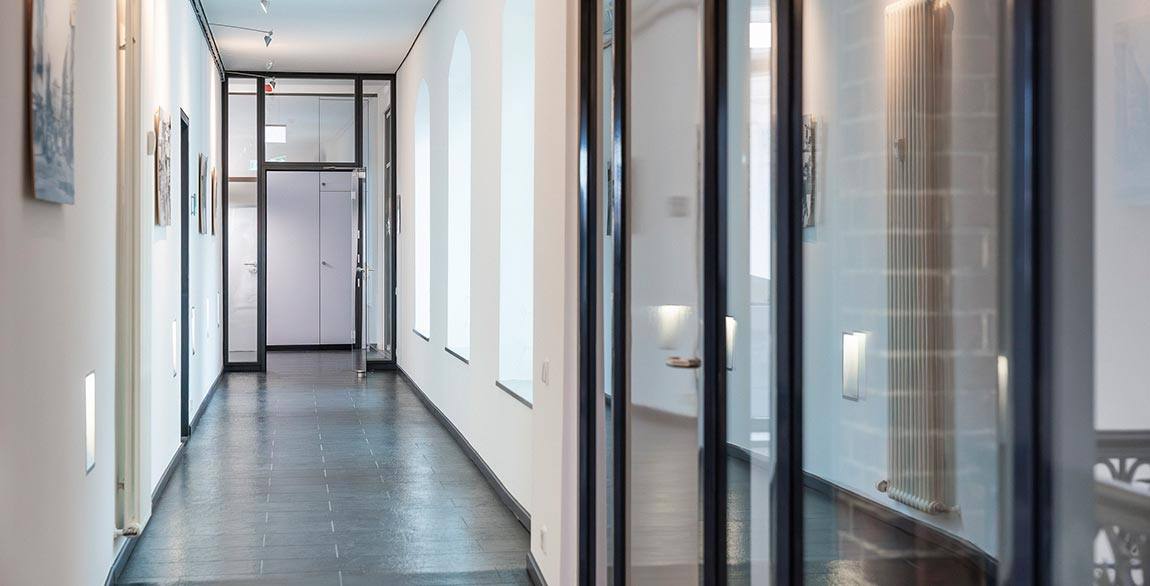 Moderner, heller Flur mit großen Fenstern und gläsernen Türen, dunklem Boden und weißen Wänden.